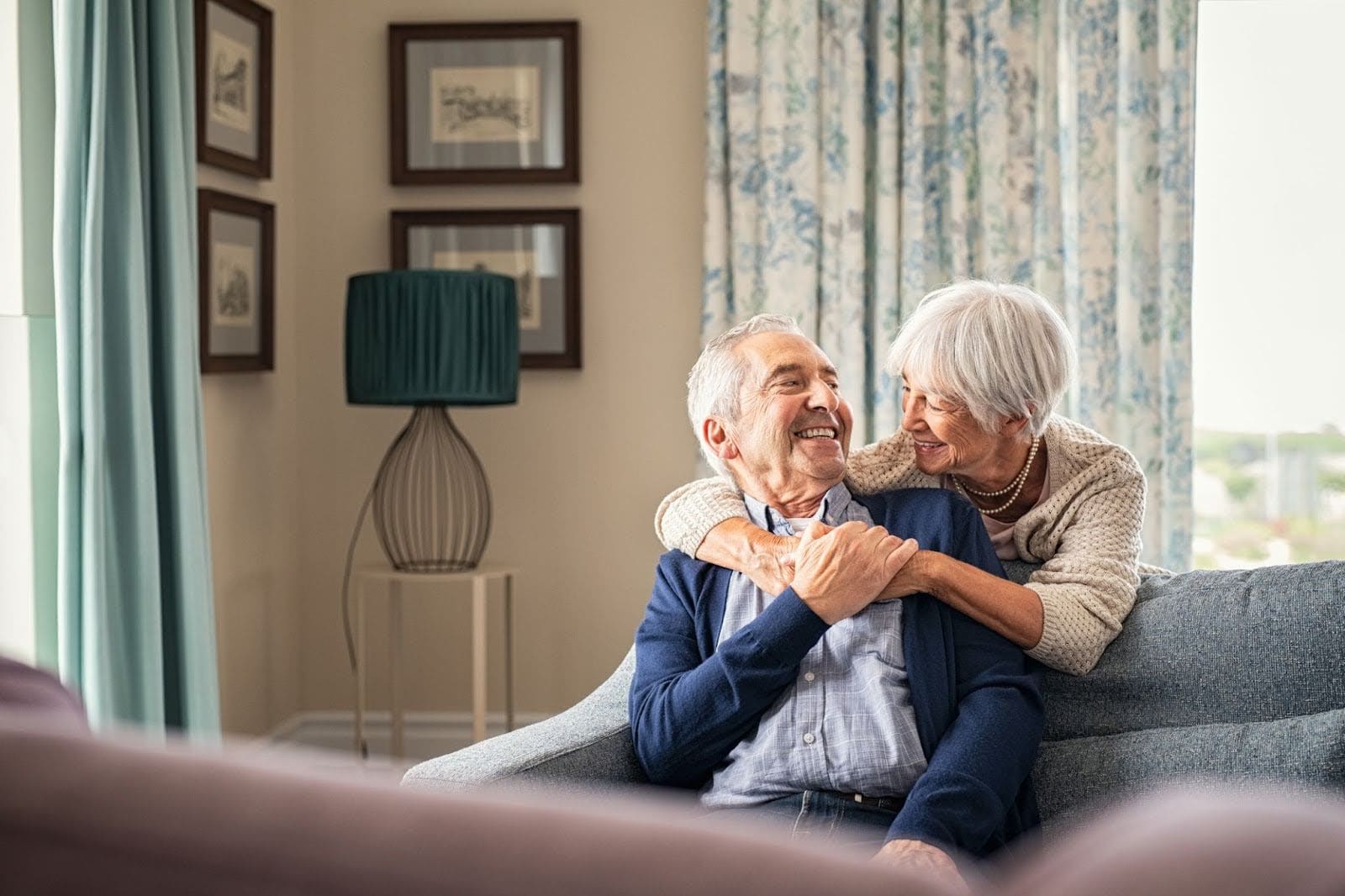 Older woman hugging her partner from behind while seated on a couch, capturing warmth and companionship at The Merion.
