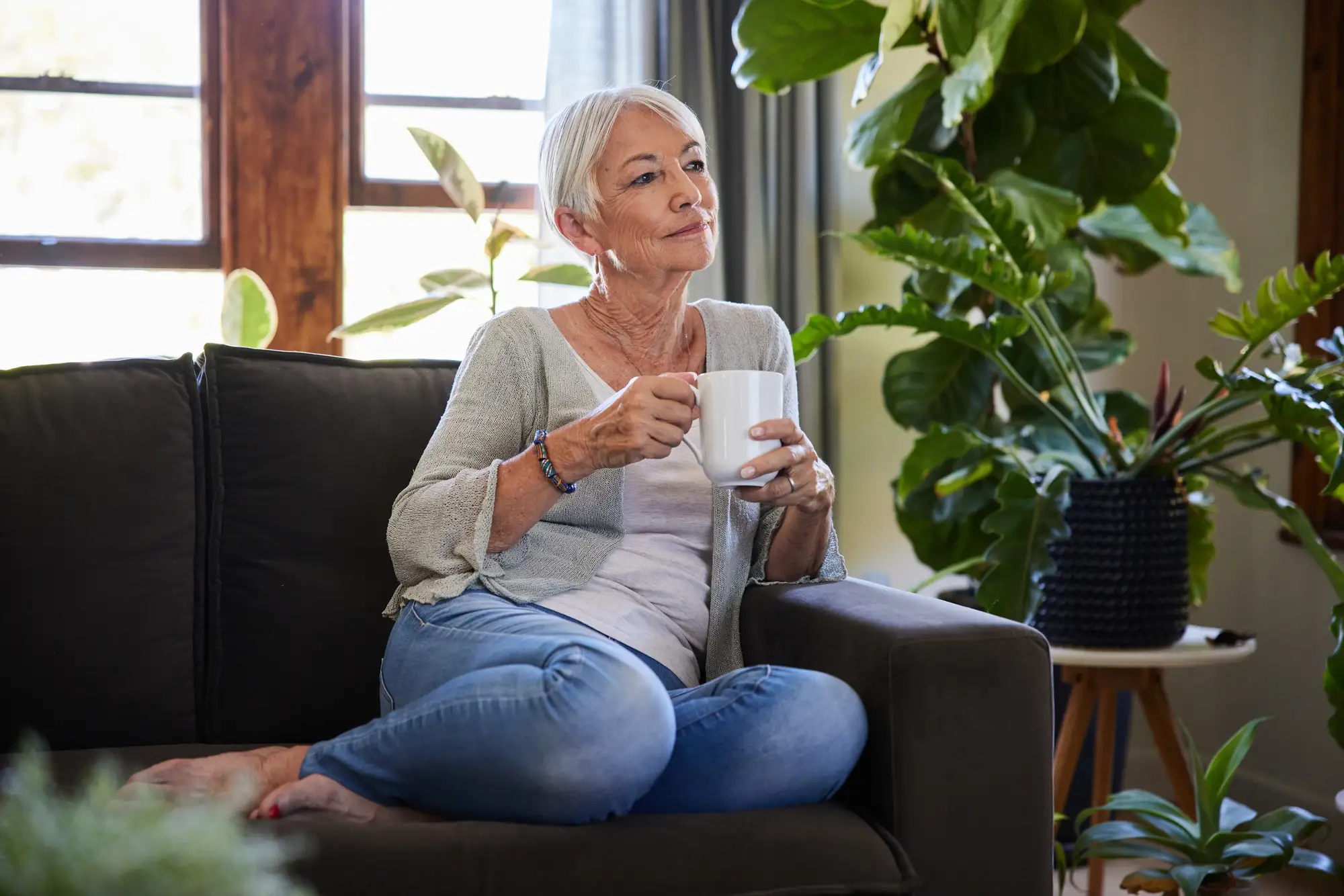 Senior woman drinking a cup of coffee and looking lost in thought while sitting on a sofa in her living room in her one-bedroom apartment